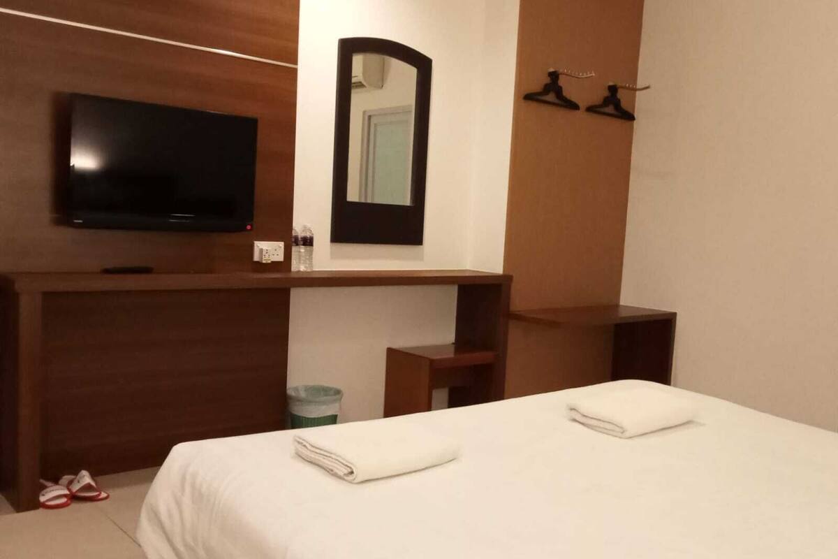 A hotel room with a bed, a TV mounted on a wooden panel, a mirror, and a shelf with two black hangers.