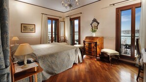 A hotel room with a bed, a desk, a chair, a dresser, a chandelier, and a view of the city. - La Calcina Suites (Venice)