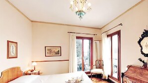 A hotel room with a large bed, a chandelier, a wooden dresser, and a balcony with a chair. - La Calcina Suites (Venice)