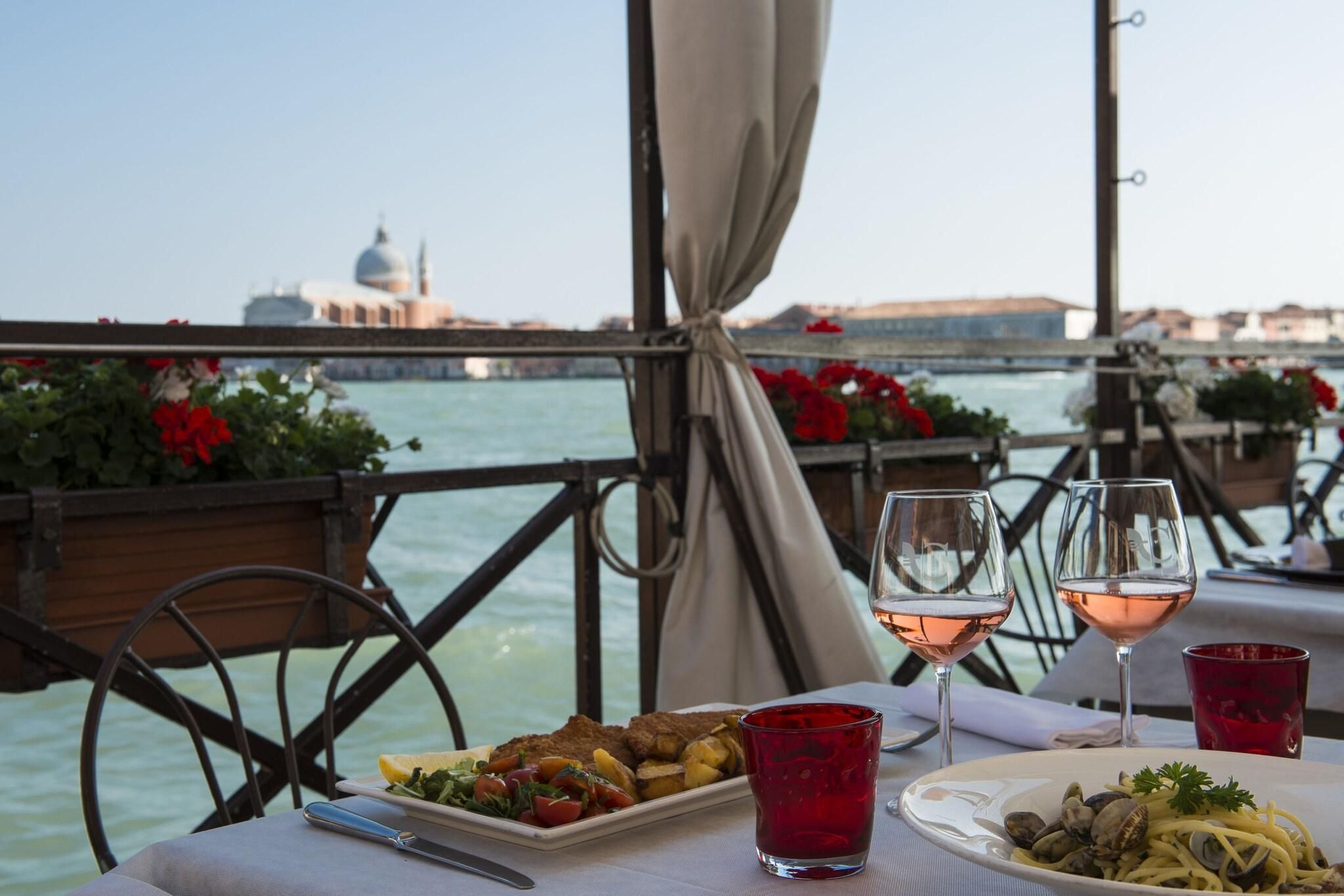 A table with plates of food and wine glasses, overlooking a body of water with a bridge and buildings in the background.