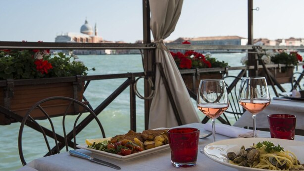 A table with plates of food and wine glasses, overlooking a body of water with a bridge and buildings in the background. - La Calcina Suites (Venice)