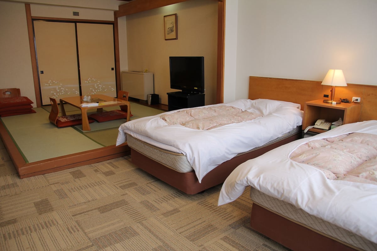 A hotel room with two beds, a television, a bedside table with a lamp, and a futon area with cushions.