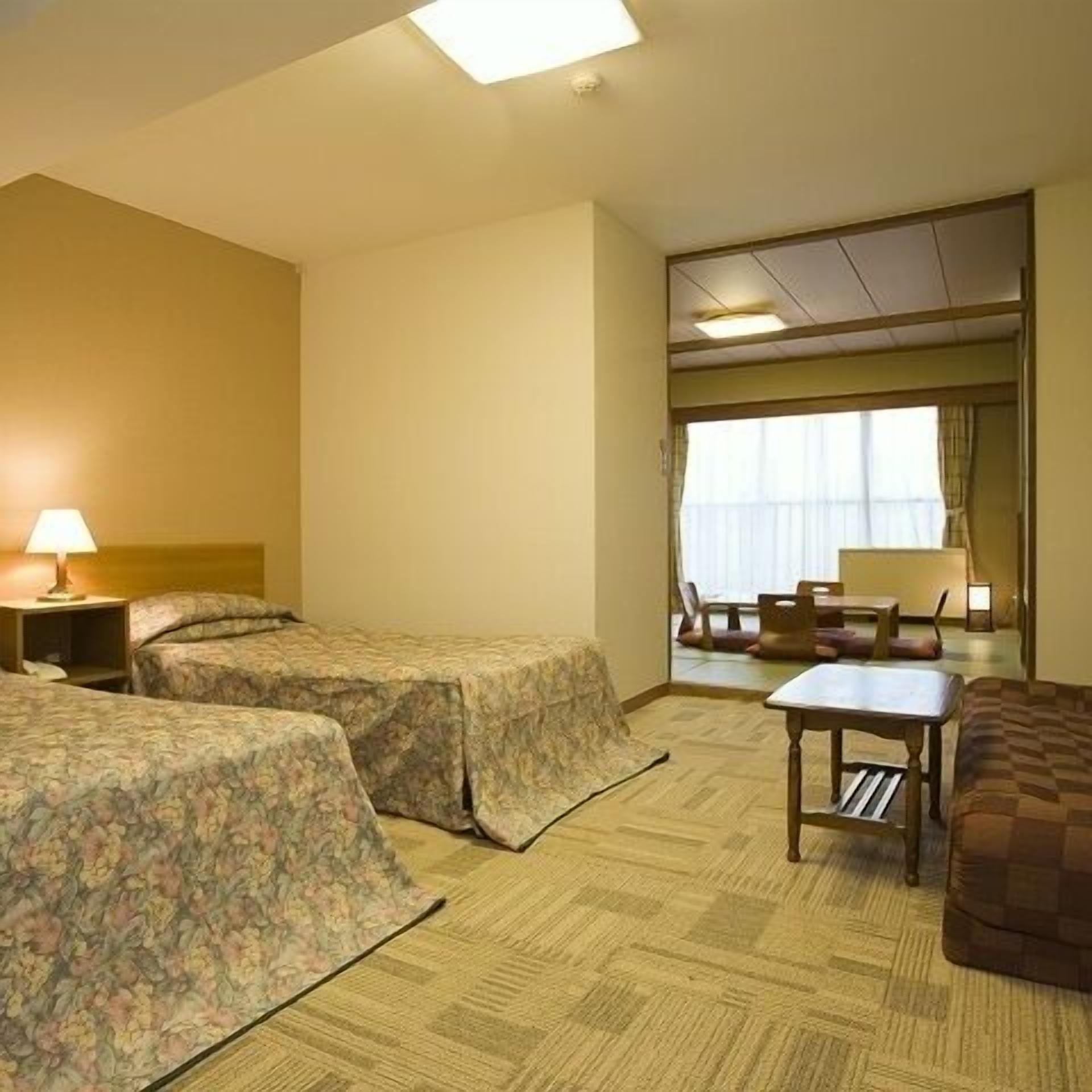 A hotel room with two beds, a small table, a chair, and a window with curtains.