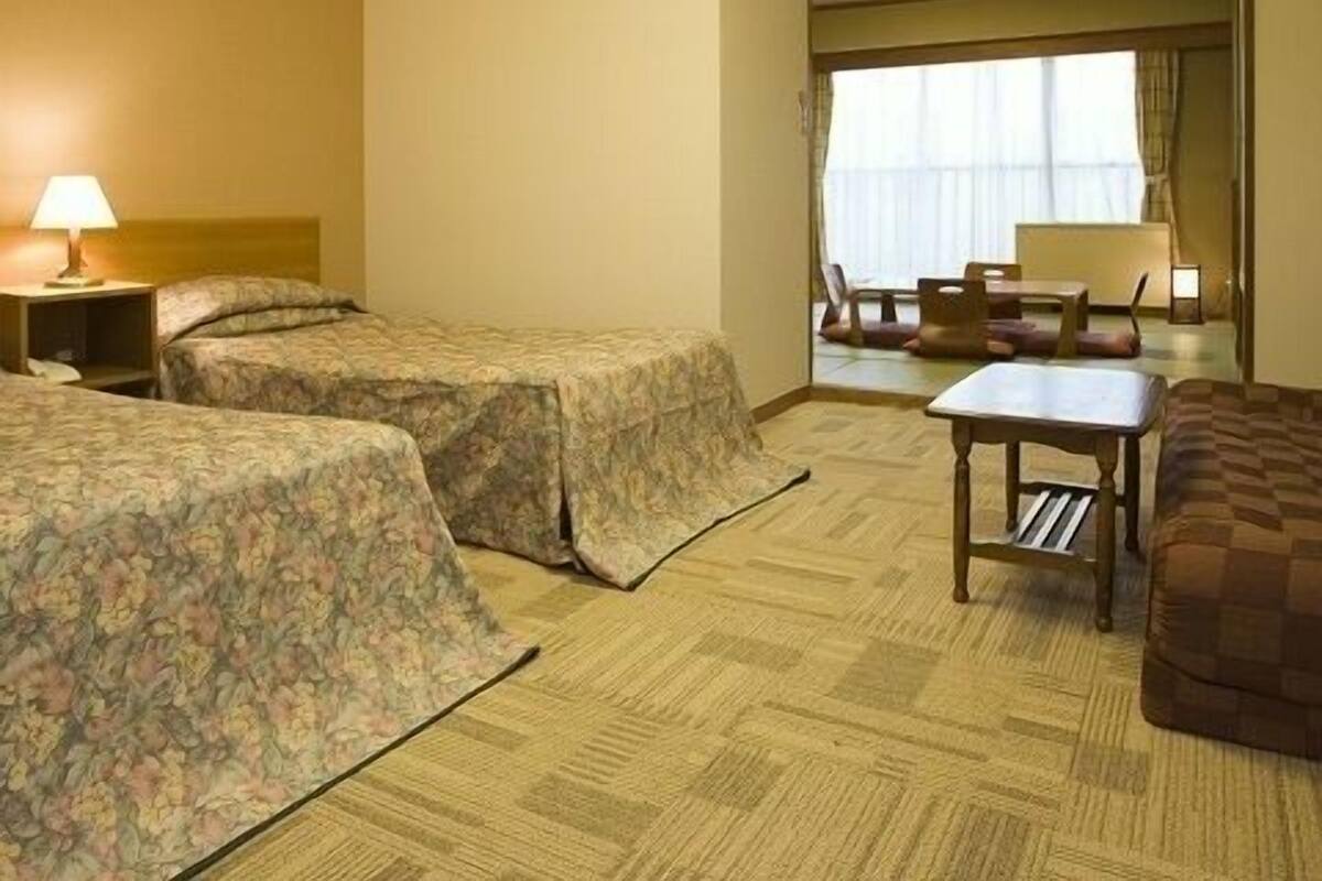 A hotel room with two beds, a small table, a chair, and a window with curtains.