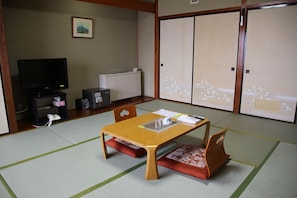 Una habitación japonesa tradicional con suelo de tatami, una mesa baja con cojines, un televisor y puertas correderas.