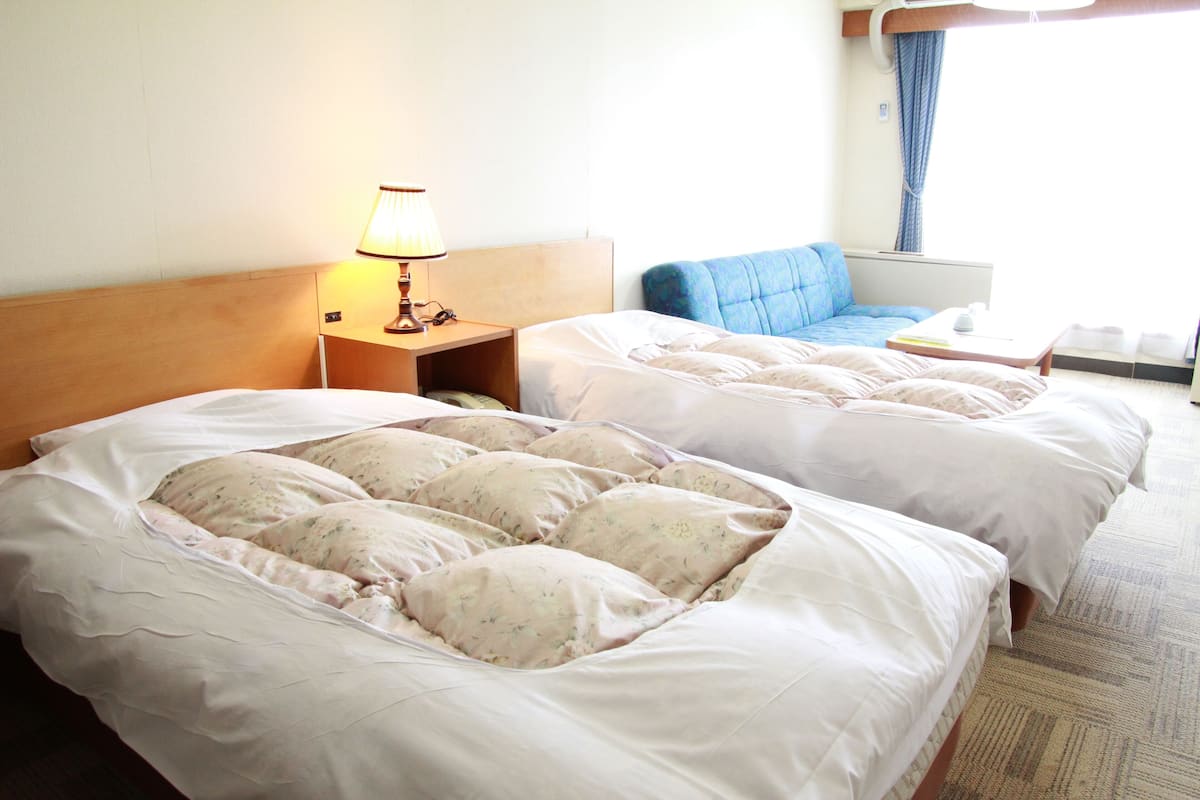 A hotel room with two beds, a nightstand with a lamp, a blue chair, and a small table with a book and a cup.