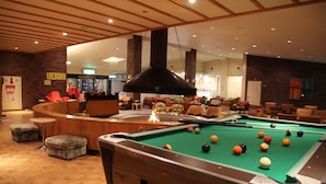 Mesa de bilhar em um saguão de hotel, com lareira, área de estar e balcão de recepção.