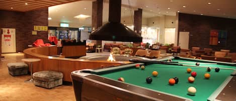 A pool table in a hotel lobby with a fire feature, seating area, and a reception desk.
