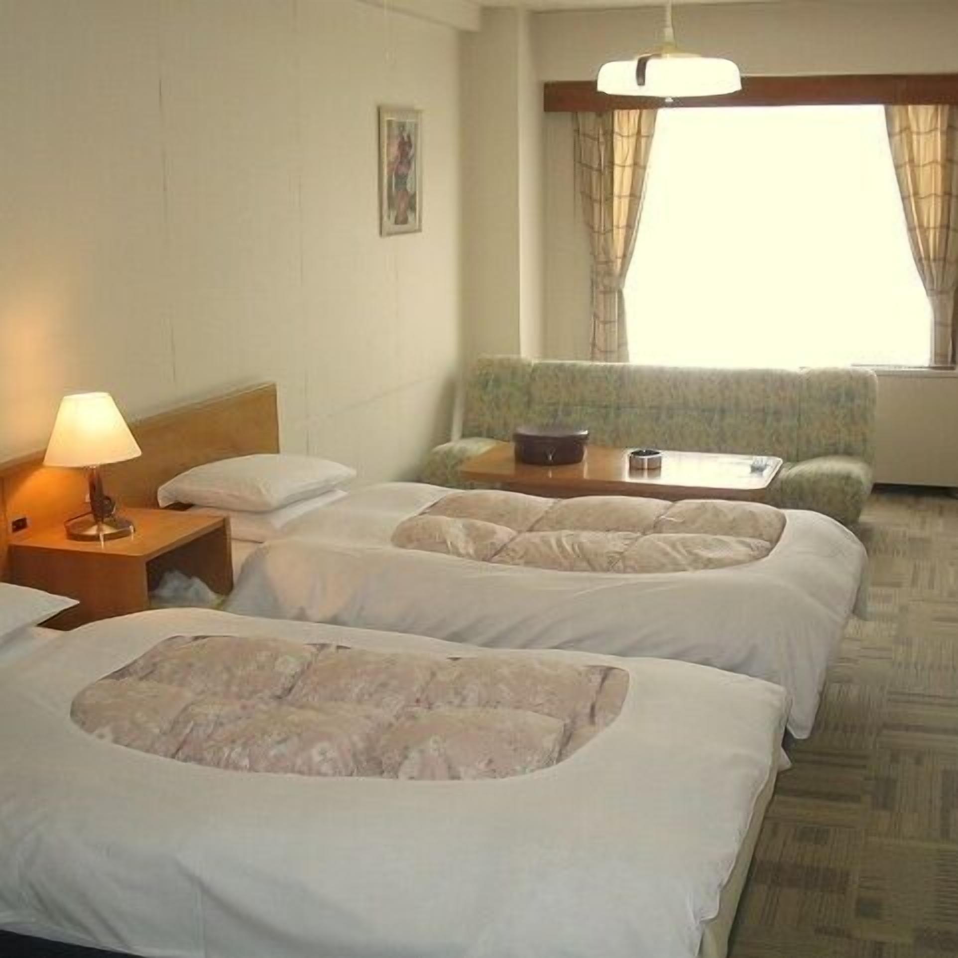 A hotel room with two beds, a bedside table with a lamp, a small table, a sofa, and a window with curtains.