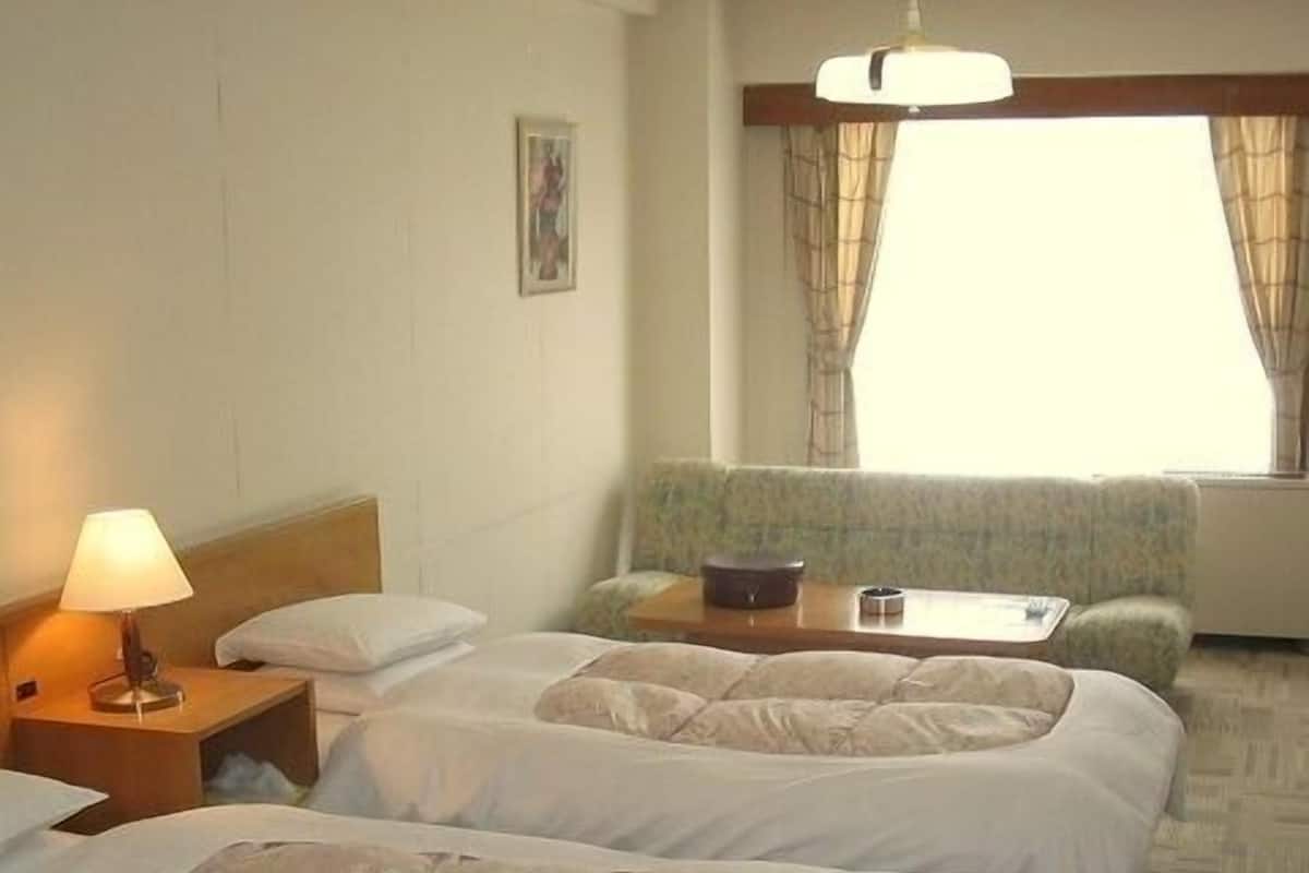 Quarto de hotel com duas camas, uma mesa de cabeceira com abajur, uma mesinha, um sofá e uma janela com cortinas.