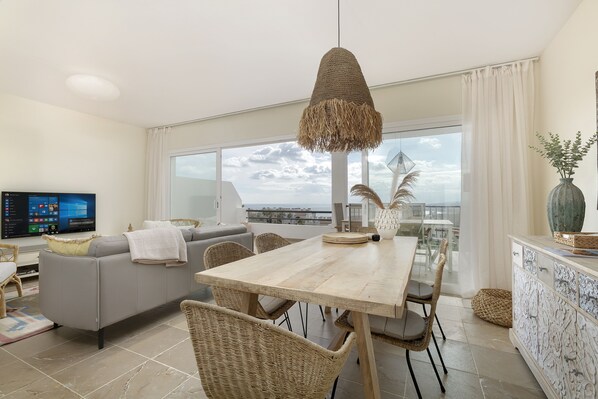 A modern living room with a grey sofa, a wooden dining table, wicker chairs, and a large window offering a city view.