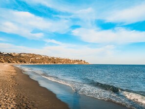 A beach with a clear sky, calm sea, and a hilly landscape in the background.
