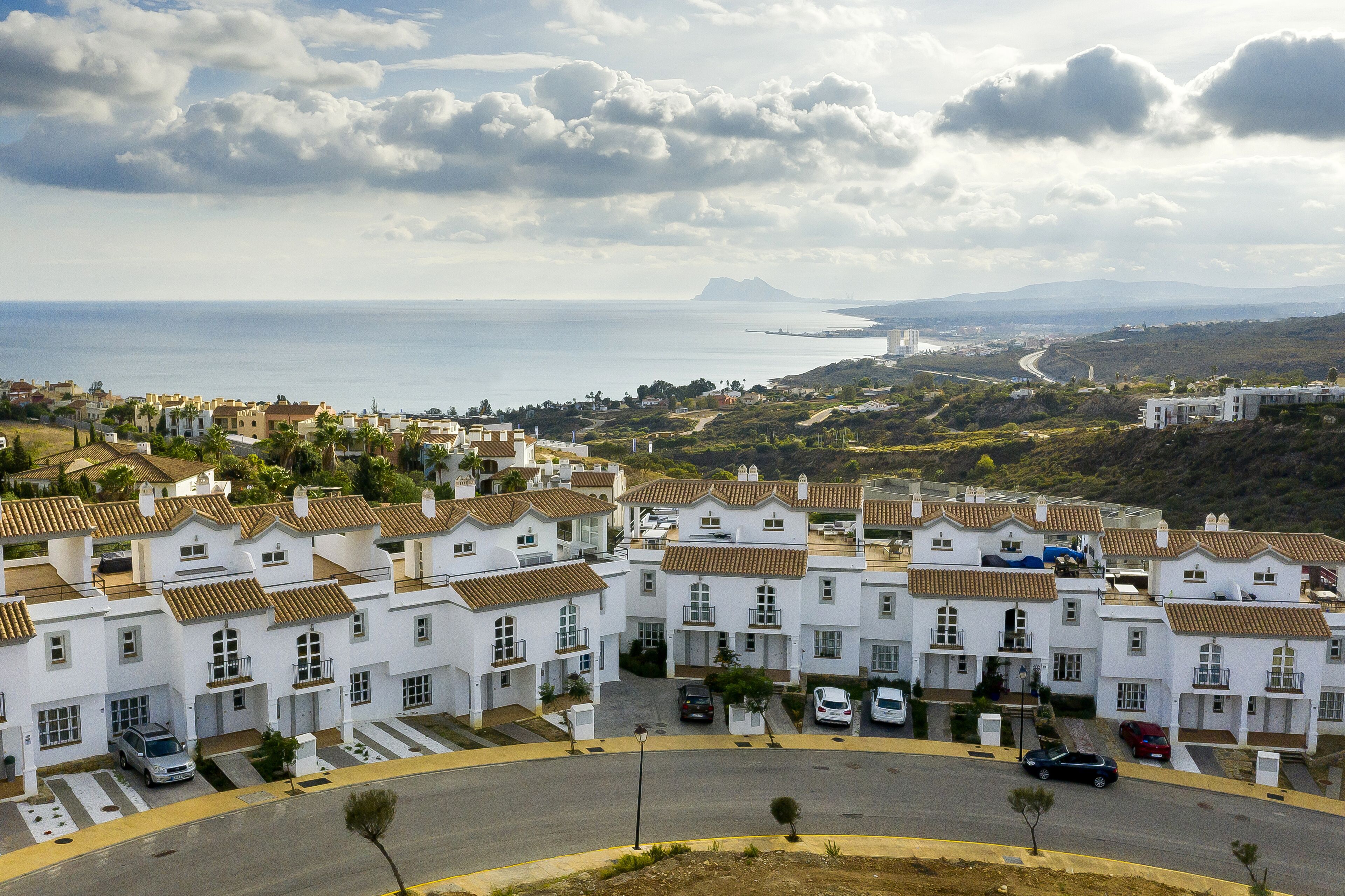 A coastal residential area with white buildings, a road, and a view of the sea and distant mountains.