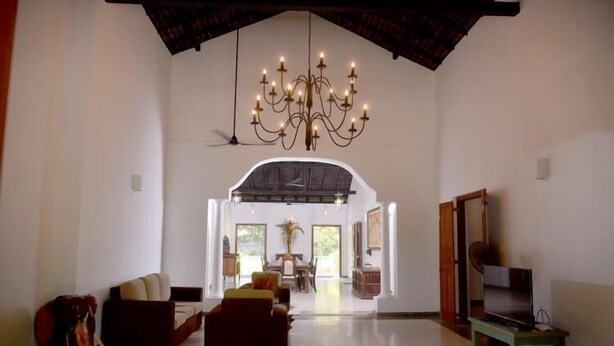 A spacious living room with a high ceiling, a chandelier, a sofa, a television, and a dining area in the background. - Ayurveda Villa 386 (Balapitiya)