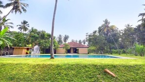 A swimming pool with a palm tree in the foreground, surrounded by lush greenery and a building in the background. - Ayurveda Villa 386 (Balapitiya)
