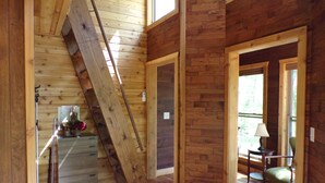 Een houten interieur met een schuine plafond, houten wanden, een trap, een eethoek met een tafel en stoelen, en een raam dat natuurlijk licht laat binnen.