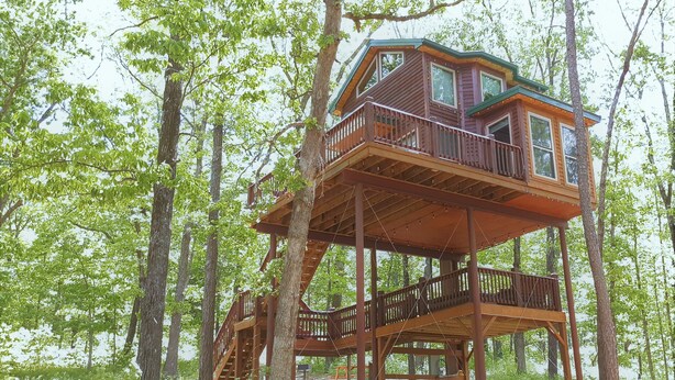 Een houten boomhut met meerdere niveaus, gebouwd rond een centrale boom, met een balkon en trap.