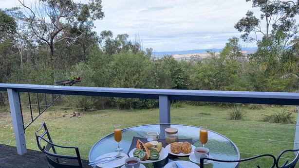 Una mesa de vidrio redonda con una comida servida, que incluye platos, cubiertos y bebidas, en una terraza con vistas a un paisaje natural.