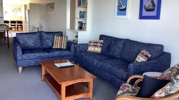 A living room with two blue sofas, a wooden coffee table, and a wicker chair. There is a bookshelf with books and framed pictures on the wall.