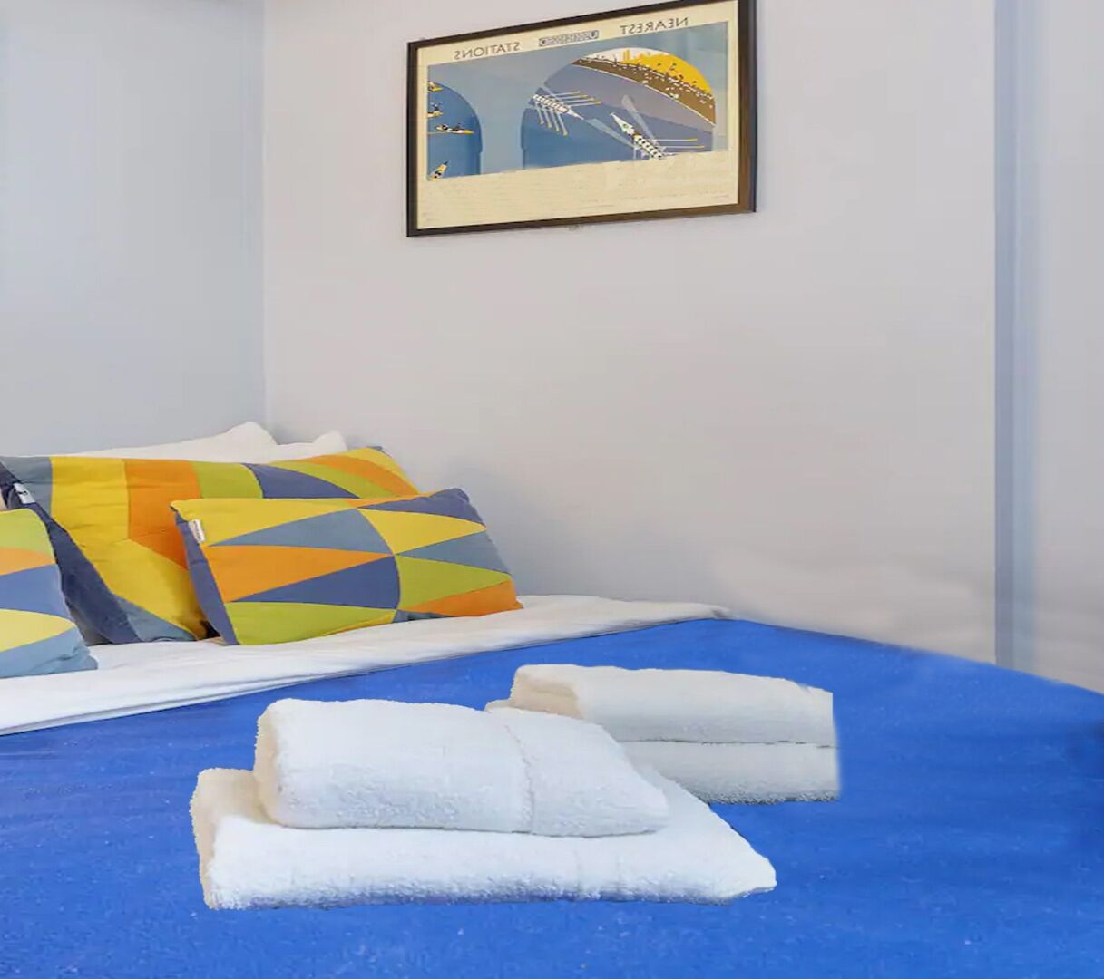A bed with a blue cover, white pillows, and a framed picture on the wall.