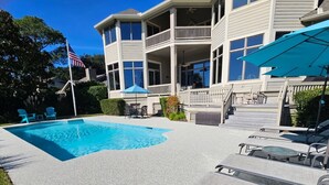 Una piscina con sillones de descanso y sombrillas, rodeada por un edificio de dos pisos con balcones.