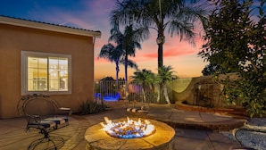 A patio with a fire pit, seating, and a view of palm trees against a sunset sky.