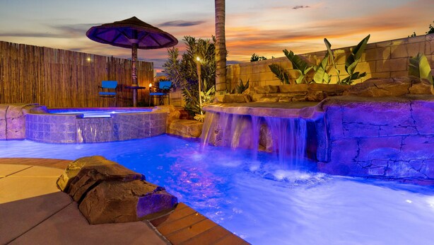A heated outdoor pool with a waterfall feature, surrounded by rocks and tropical plants, and a wooden fence in the background.