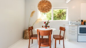 A dining area with a round table, four wooden chairs, a woven light fixture, a vase with flowers, and a window with a view of greenery.