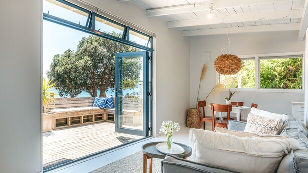A modern living room with a large sliding glass door leading to an outdoor deck, a grey sofa, a small round table, and a vase with flowers.