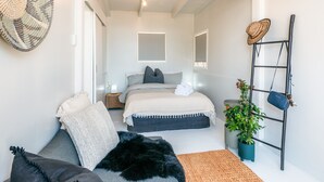 A modern bedroom with a bed, a sofa, a small table, a plant, and a ladder with a hat and bag.