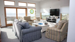 A spacious living room with a large TV, comfortable seating, and a view of the ocean through large windows.