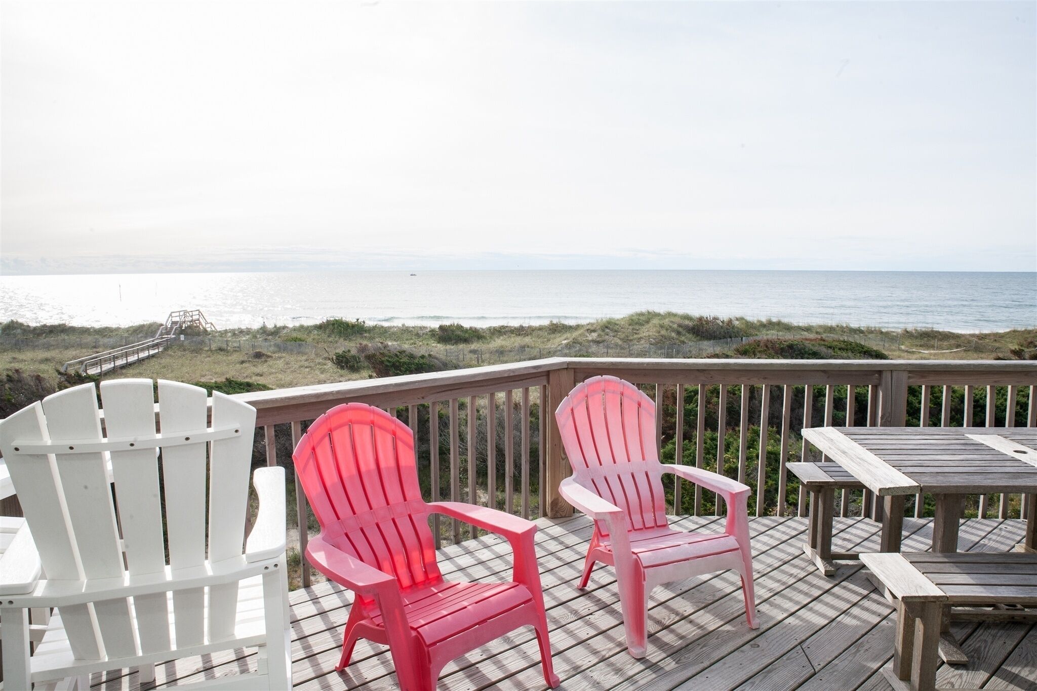 A deck with Adirondack chairs and a wooden table overlooking a beach.