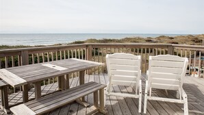 A wooden deck with a table and two white chairs, overlooking a beach with dunes and a clear sky.