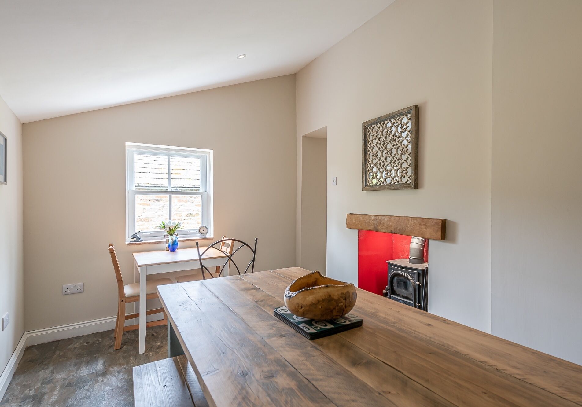A dining area with a wooden table, chairs, a vase with flowers, a fireplace, and a window with blinds.