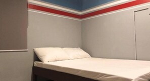 A small, single-bed room with a plain white mattress and pillows, a wooden headboard, and a plain wall with a red and blue stripe at the top. - Homeland Tourister's Apartelle Boracay (Boracay Island)