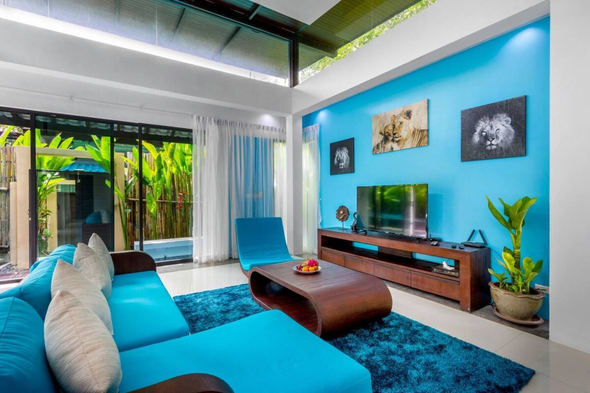 Ein modernes Wohnzimmer mit einem blauen Sofa, einem Couchtisch aus Holz und einem Fernseher auf einem Holzständer. An der Wand hängen zwei Gemälde, und es steht ein Blumentopf bereit. Ein Schiebetürfenster führt auf einen Balkon mit Pflanzen.