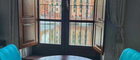 A room with a round table and two turquoise chairs, a window with a view of buildings and a canal.