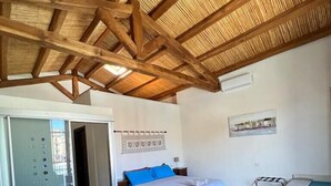 A spacious room with a wooden ceiling, a bed with blue pillows, a dining table with chairs, and a wall-mounted air conditioning unit.