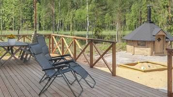Puinen terassi, jossa on metallinen grilli, pieni puinen rakennelma ja taustalla metsä.
