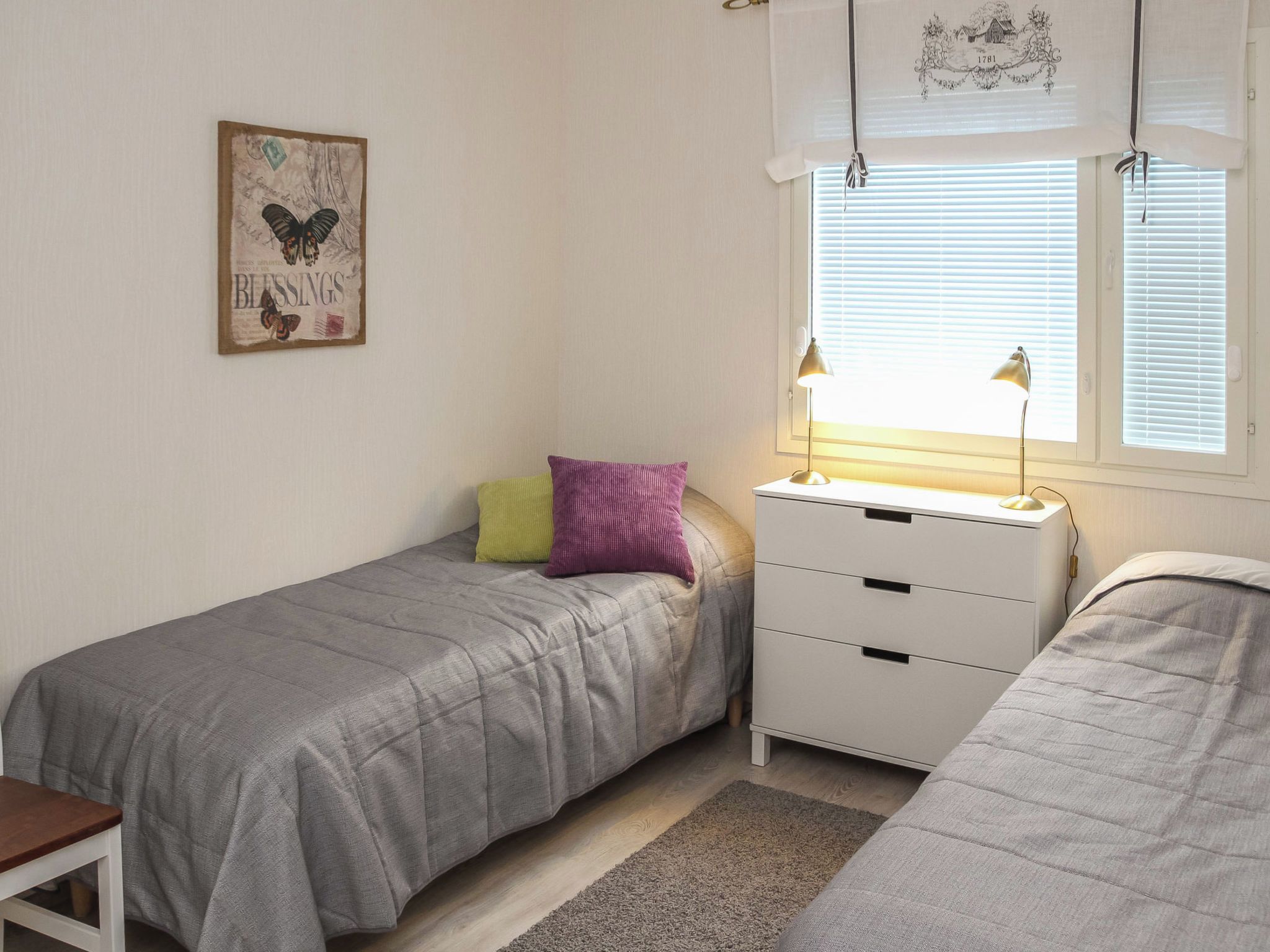 A bedroom with two single beds, a bedside table with a lamp, a window with blinds, and a wall-mounted picture.