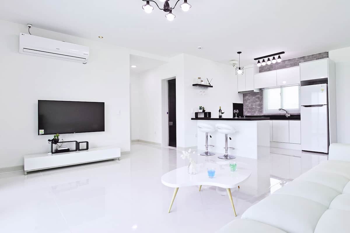 A modern living room with a flat-screen TV, a white sofa, and a dining area with two bar stools.