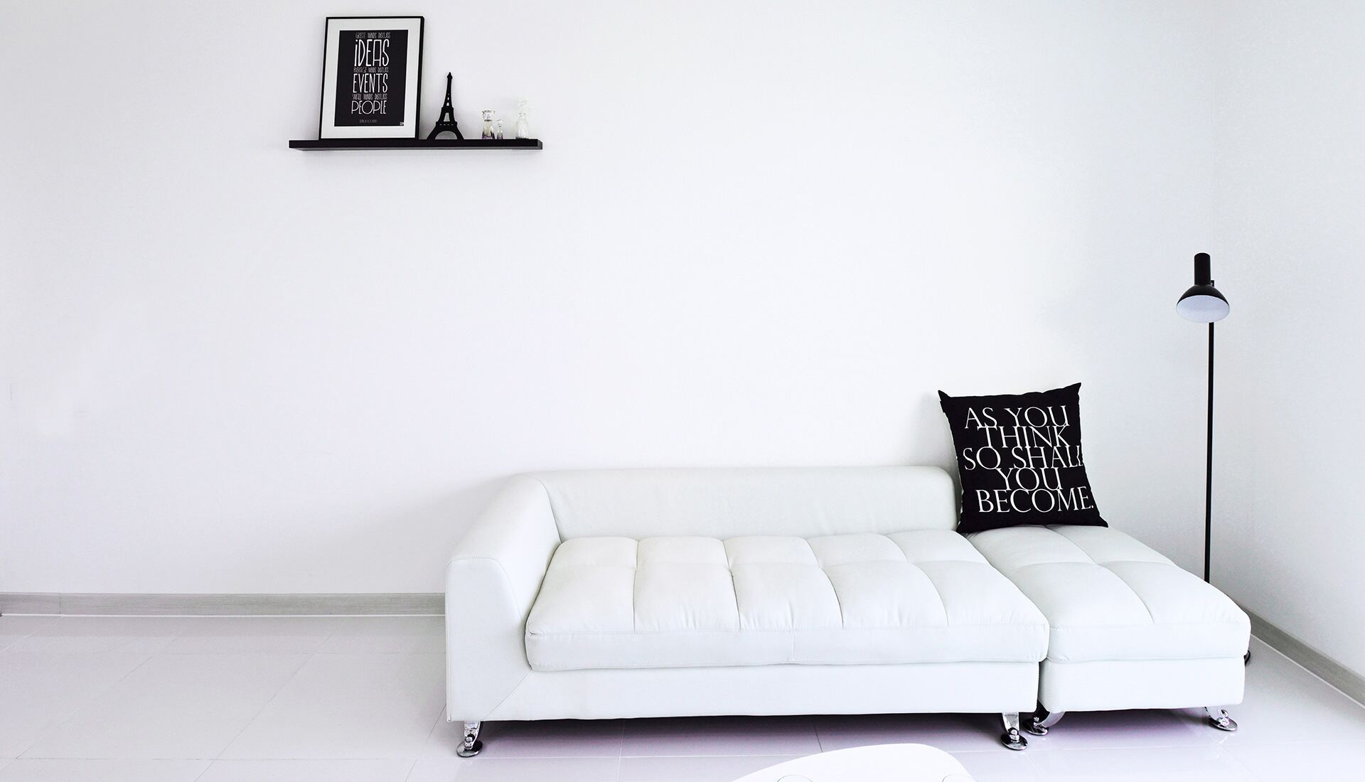 Un canapé blanc avec un coussin noir, un lampadaire et un tableau encadré sur un mur blanc.