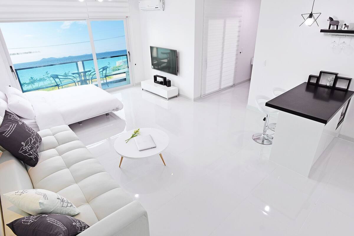 A modern living room with a sofa, a small round table, a TV, and a balcony with a view of the sea.
