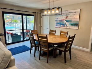 A dining area with a round wooden table and six chairs, a ceiling light fixture, and a view of a swimming pool through a sliding glass door.