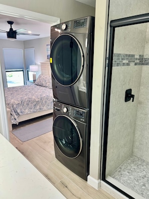 Une chambre d’hôtel compacte dotée d’un lave-linge et d’un sèche-linge à double porte, d’un lit avec une literie à motifs, d’un ventilateur de plafond et d’une vue sur l’extérieur grâce à une fenêtre équipée de stores.
