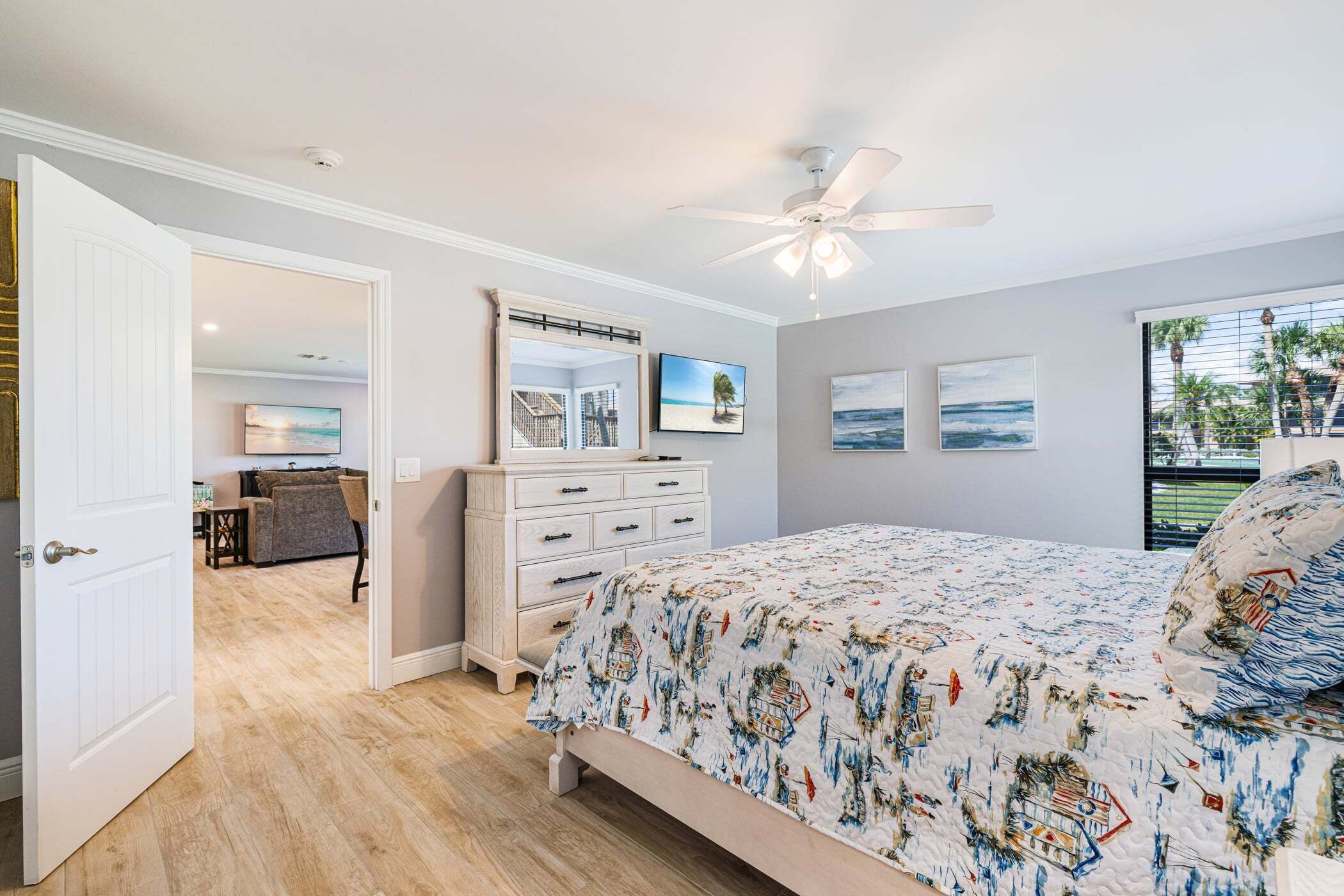 A bedroom with a bed, a dresser, a ceiling fan, and a view of the beach through a window.
