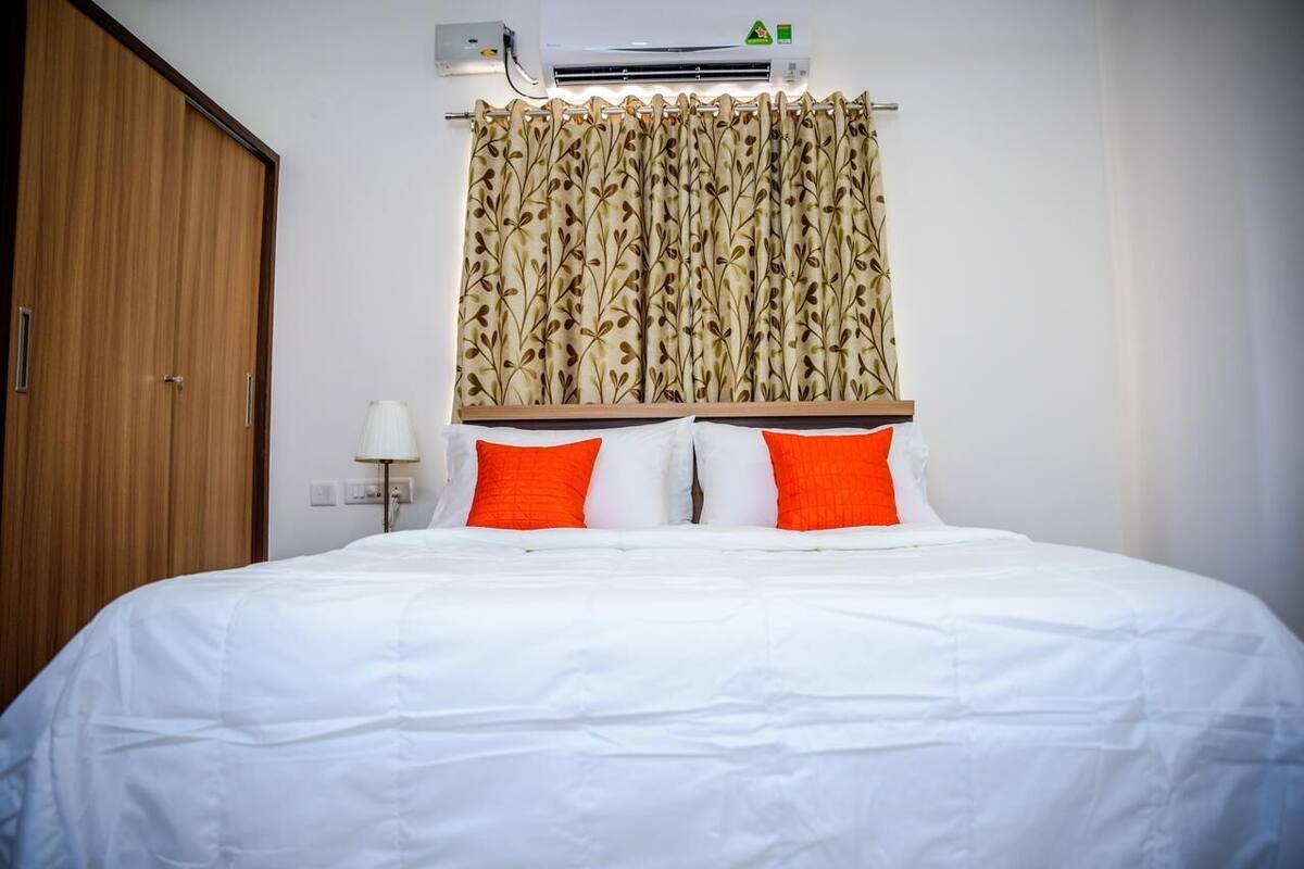 A hotel room with a bed, white bedding, two orange pillows, a wooden wardrobe, a wall-mounted air conditioner, and a curtain with a leaf pattern.