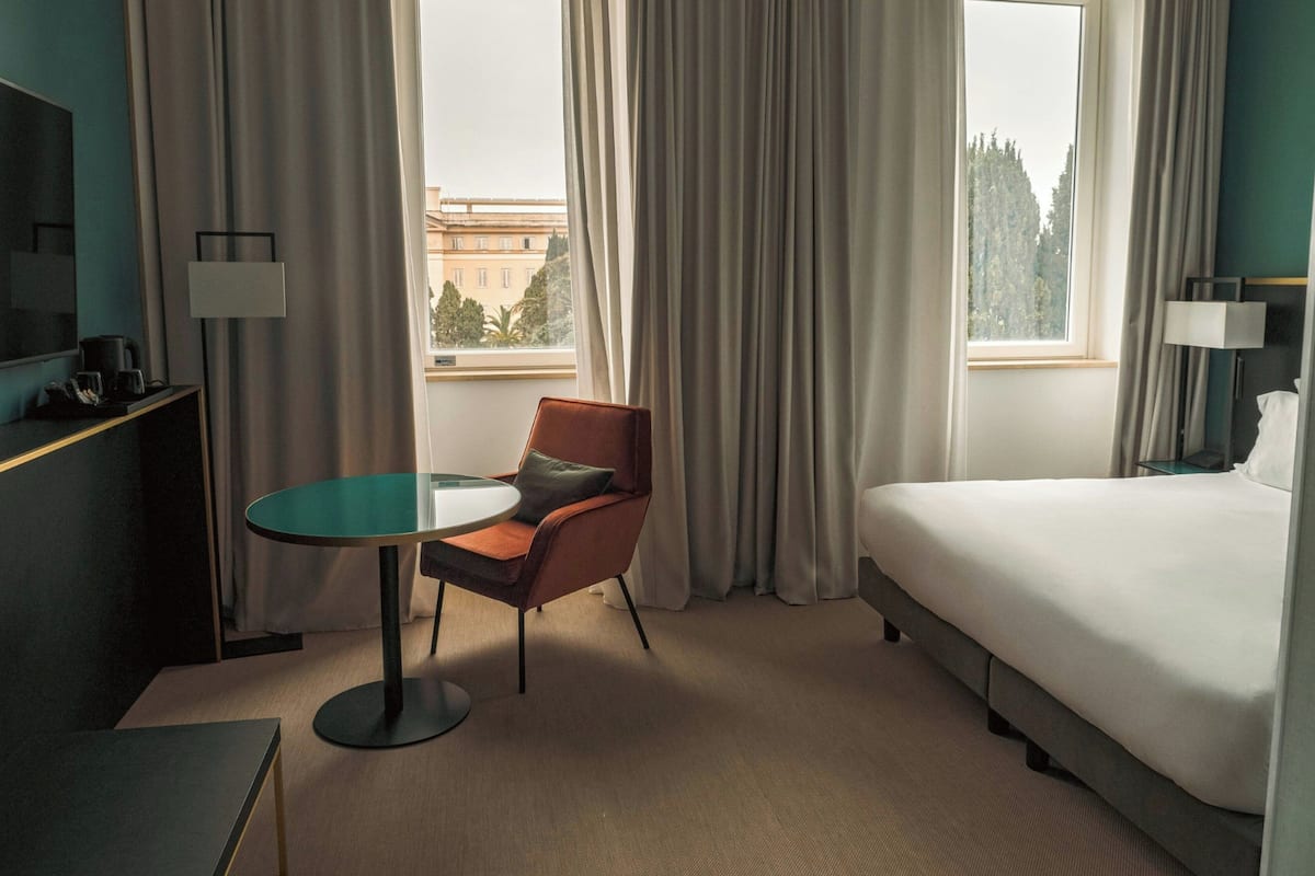 Quarto de hotel com cama, mesa redonda, cadeira, televisão e janela com cortinas.