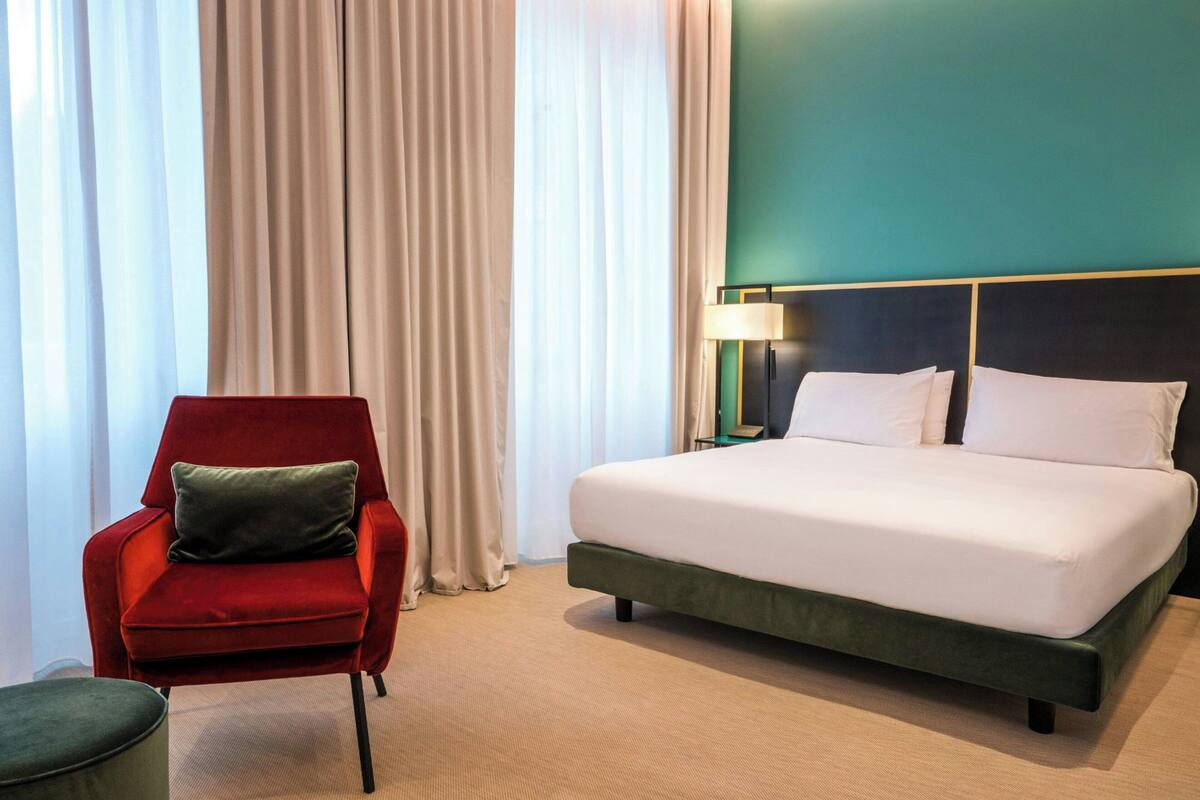 Quarto de hotel com uma cama, uma poltrona vermelha, um banquinho verde e uma mesa de cabeceira com luminária.