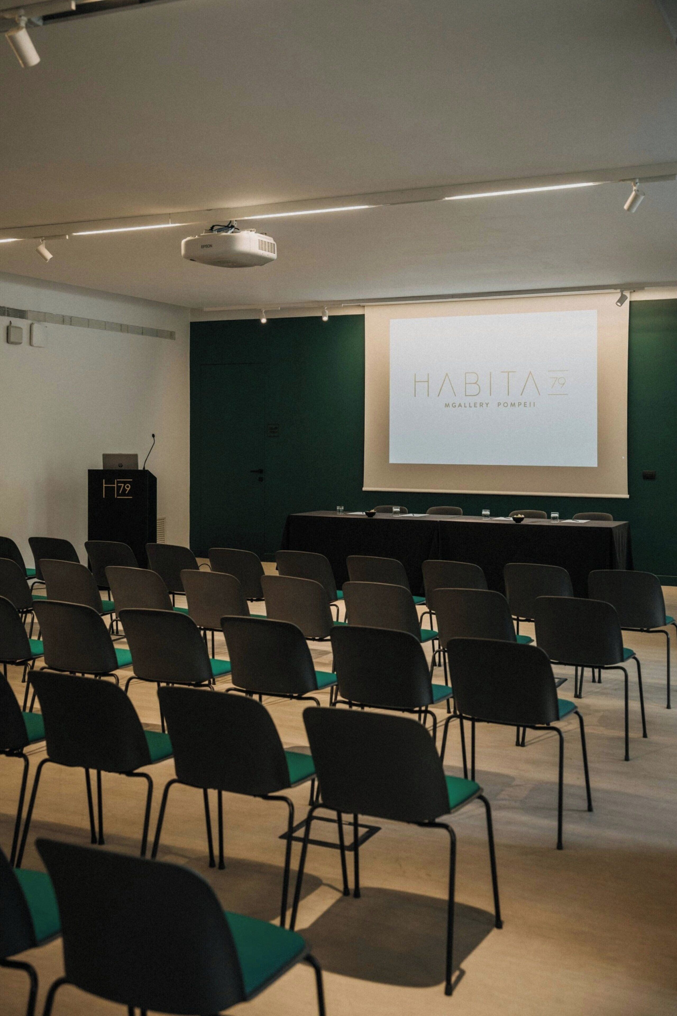 A conference room with rows of chairs, a podium, and a projection screen displaying 'HABITA'.